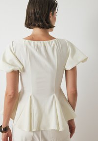 Witte peplum blouse met puffmouwen, textuurstof en een geplooid neklijn-detail. De achterkant heeft een aansluitende pasvorm die naar de peplum toelopert.