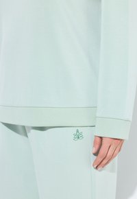 Conjunto de algodón verde claro que incluye una camiseta de manga larga con puños acanalados y pantalones a juego. Pequeño detalle del logo cerca del dobladillo.