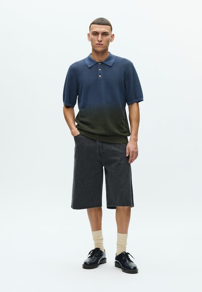 Πόλο πουκάμισο με διαβάθμιση από σκούρο μπλε σε πράσινο, με κοντά μανίκια, συνδυασμένο με σκούρο τζιν σορτς έως τα γόνατα και μαύρα παπούτσια.