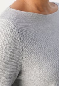 Lähikuva henkilöstä, jolla on päällään vaaleanharmaa teksturoitu neulepaita pyöreällä kaula-aukolla, ja näkyy olkapää ja ylärinta-alue.