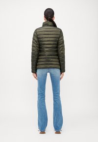 Doudoune vert olive avec un matelassage horizontal, un col montant et une texture lisse, associée à un jean bleu évasé.