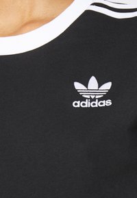 Černé tričko s bílým žebrovaným límcem, se vyšitým bílým logem Adidas a třemi bílými pruhy na rameni.