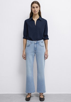 Navy button-up shirt with rolled sleeves paired with light blue flared jeans. Model wears black flats. Plain background.