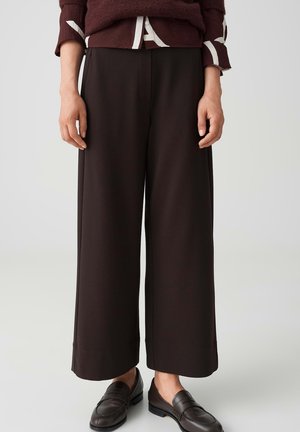 Personne portant un pantalon large marron foncé, des mocassins en cuir noir et un pull bordeaux par-dessus une chemise blanche.