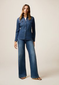 Camicia in denim blu scuro con chiusura a bottoni e tasche sul petto, abbinata a jeans a gamba larga con decorazioni in perline sui lati.