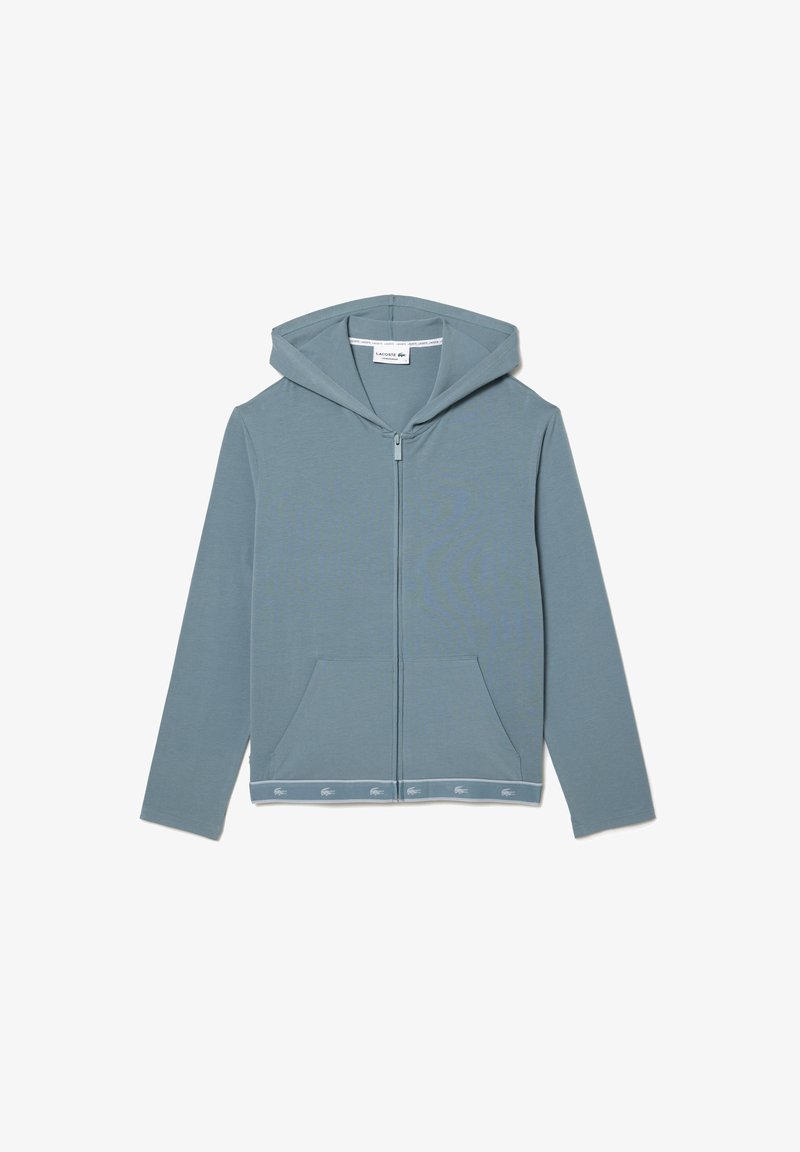 Lys blå hettejakke med glidelås laget av myk stoff. Har en frontlomme, hette og et bånd med logo ved kanten. Enkelt design.