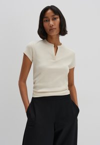 Light beige ribbed short-sleeve top with a small collar and a keyhole cutout at the neckline, paired with high-waisted black trousers.