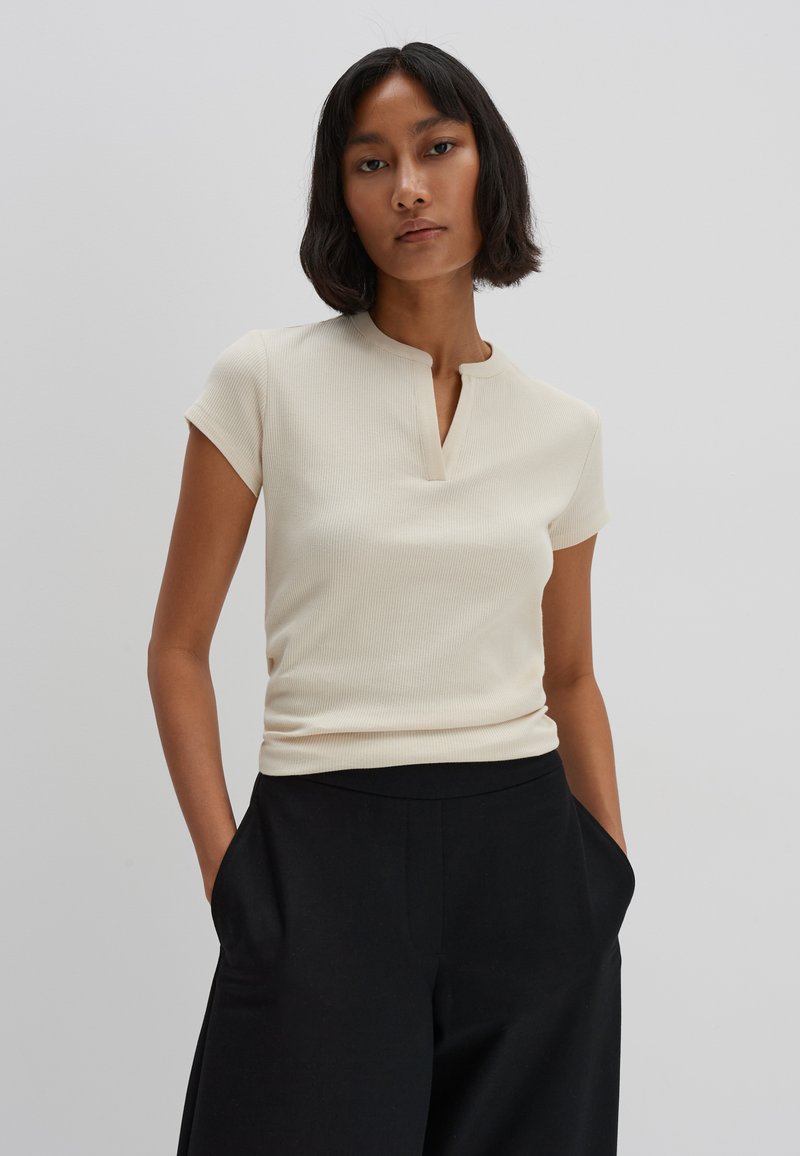 Light beige ribbed short-sleeve top with a small collar and a keyhole cutout at the neckline, paired with high-waisted black trousers.