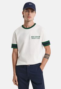 White short-sleeve T-shirt with green and navy striped accents, featuring "NEW HAVEN SPORTS CLUB" print on the left chest. Navy cap.