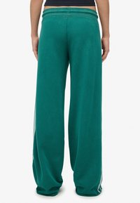 Persoon in een teal brede broek en een zwart cropped top, gezien van achteren tegen een effen witte achtergrond.
