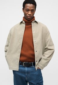 Veste en velours côtelé vert clair avec des boutons-pression argentés, superposée sur un col roulé côtelé marron, associée à un jean bleu foncé et une ceinture noire.