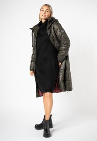Olivengrüner Puffer-Jacke mit Kapuze, kombiniert mit einem langen schwarzen Strickkleid. Schwarze Stiefeletten vollenden den Look. Glatte Textur und praktisches Design.