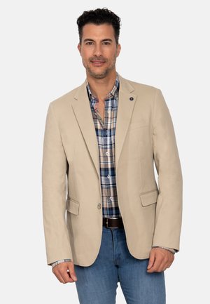 Blazer beige de tela ligera con solapas con muesca, bolsillos laterales y un pequeño botón decorativo. Llevado sobre una camisa a cuadros.
