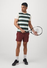 Mees, kellel on seljas triibuline tennisepaita, maroonid lühikesed püksid, valged sokid ja mustad kingad, hoiab tennisereketit ning vaatab küljele ühetoonilise tausta ees.