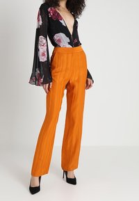 Blusa nera a maniche lunghe con stampa floreale e scollatura a V profonda; abbinata a pantaloni arancioni a vita alta con texture e tacchi alti neri.
