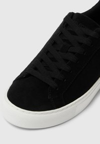 Sneaker in suede nera con lacci piatti neri, suola in gomma bianca e punta testurizzata. Superficie liscia e design minimalista.