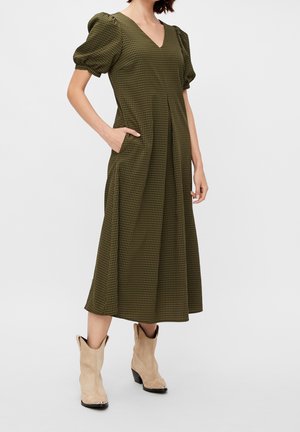 Mujer con vestido midi de cuadros verdes y negros con mangas abullonadas y botas cowboy color beige, de pie con una mano en el bolsillo.