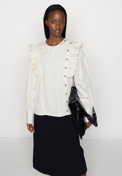 Blusa cremosa de mangas longas com detalhe de ombreiras em babados e fecho com botões. Combinada com uma saia preta e segurando uma bolsa de couro preta.