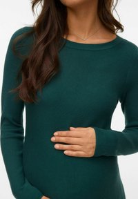 Stickad, smaragdgrön tröja med ribbad textur, båthals och långa ärmar. Hand placerad varsamt på midjan.