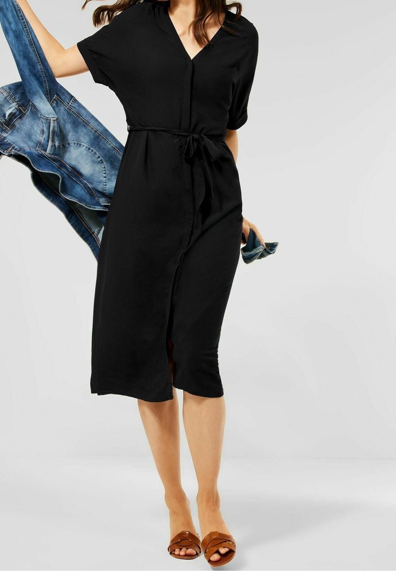 Femme portant une robe midi noire à manches courtes et taille cintrée avec un lien, tenant une veste en jean bleue et une pochette, portant des sandales marron.