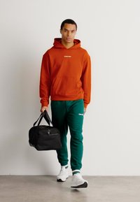 Felpa arancione con cappuccio e tasca frontale, pantaloni della tuta verdi e borsa a tracolla nera. Il modello indossa sneaker bianche. Design semplice.