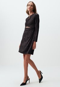 Zwarte jurk met lange mouwen, tailleaccentuering en uitsnijdetails. Heeft een subtiel bloemenpatroon en een gladde textuur. Gecombineerd met zwarte hakken.