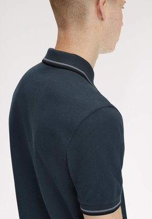 Uomo con capelli corti che indossa una polo blu scuro con colletto e bordi delle maniche a righe, mostrato di spalle e di lato.