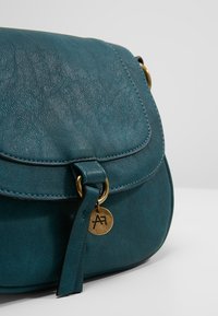 Torba na ramię z turkusowej skóry z klapką, z metalowym zapięciem i złotym zawieszkiem z inicjałami. Gładka faktura i zaokrąglony kształt.