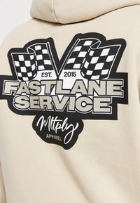 Sweat-shirt beige avec un grand graphisme noir comprenant des drapeaux à damier, "FASTLANE SERVICE" et "Mltply APPAREL." Matière douce en coton.