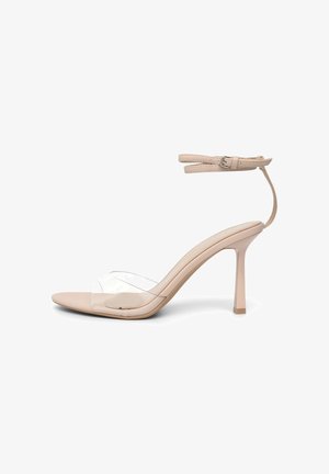 Sandale à talon beige avec une bride transparente sur les orteils et des doubles brides à la cheville avec fermeture à boucle sur fond blanc.