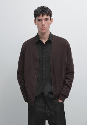 Bruine cardigan van zachte stof met een v-hals en knoopsluiting, gelaagd over een donker geruit shirt, aangevuld met een donkere jeans.