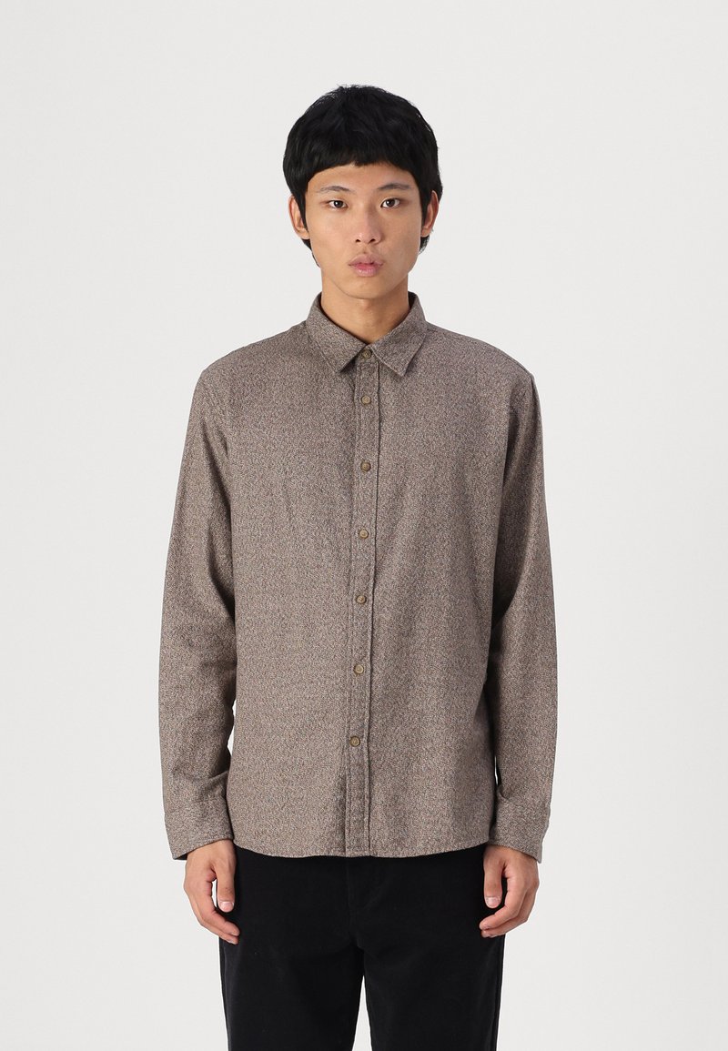 Καφέ πλεκτό πουκάμισο με γιακά σε σχήμα φιόγκου και μακριά μανίκια, με διακριτό σχέδιο και αντιθετικούς κόμπους. Φορεμένο με μαύρο παντελόνι από κάτω.