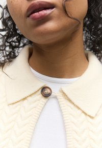 Nærbillede af en person iført en cremefarvet strikket cardigan med et tekstureret mønster og en stor brun knap over en hvid ribbet top.