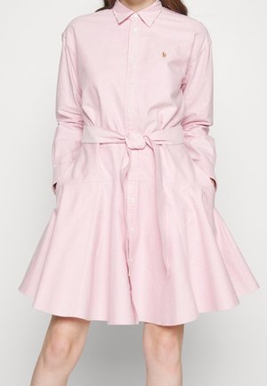 Femme portant une robe chemise rose clair avec manches longues, ceinture nouée à la taille et jupe évasée, debout les mains dans les poches.