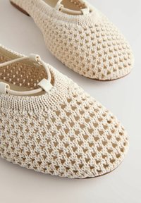 Des chaussures plates tricotées de couleur crème présentent un motif de tissage ouvert, un bout arrondi et une fine lanière de cheville avec une petite fermeture.