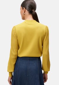 Pull en maille jaune moutarde avec des manches bouffantes à côtes. Design lisse au dos, porté avec une jupe en denim.