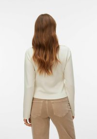 Ivoorkleurige top met lange mouwen en een afgeronde zoom, gecombineerd met lichtbeige, hooggetailleerde jeans. Soepele stof en eenvoudig ontwerp. Haar gestyled in losse golven.