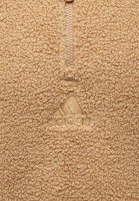 Pješački pulover od pješčenjaka s patentnim zatvaračem na vratu. Ima teksturiranu površinu i utisnutu Adidas logotip u donjem kutu.