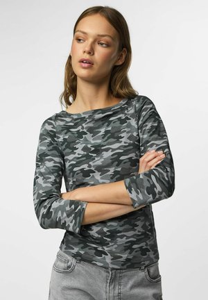 Jonge vrouw met lichtbruin haar die een grijs-zwart camouflage langarmshirt en lichtgrijze spijkerbroek draagt, armen gekruist.