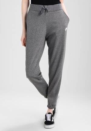 Osoba nosząca szare spodnie dresowe Nike z sznurkiem w talii, bocznymi kieszeniami oraz czarne trampki z białymi sznurowadłami i podeszwami.