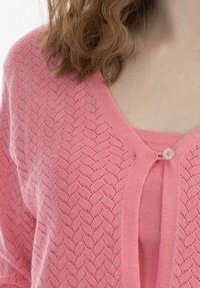 Rosa, stickad cardigan med ett texturerat, chevronmönster och en knappstängning. Underneath finns en enfärgad rosa topp.