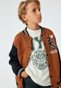 Brun og sort varsity-jakke med stribede manchetter, der har knapper og et print på fronten. Båret over en hvid t-shirt med grøn grafik.