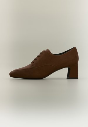 Zapato de vestir para mujer en ante marrón con tacón bajo, cordones y puntera cuadrada, mostrado de lado sobre un fondo neutro.