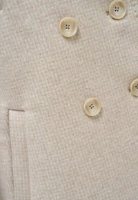 Beige stoffen jas met textuur, voorzien van vier lichtgekleurde knopen en een horizontale zak aan de linkerkant.