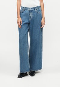 Άτομο που φοράει μπλε τζιν με φαρδιά πόδια και ψηλή μέση, καθώς και μαύρα παπούτσια με αιχμηρή μύτη, που στέκεται μπροστά σε λευκό φόντο.