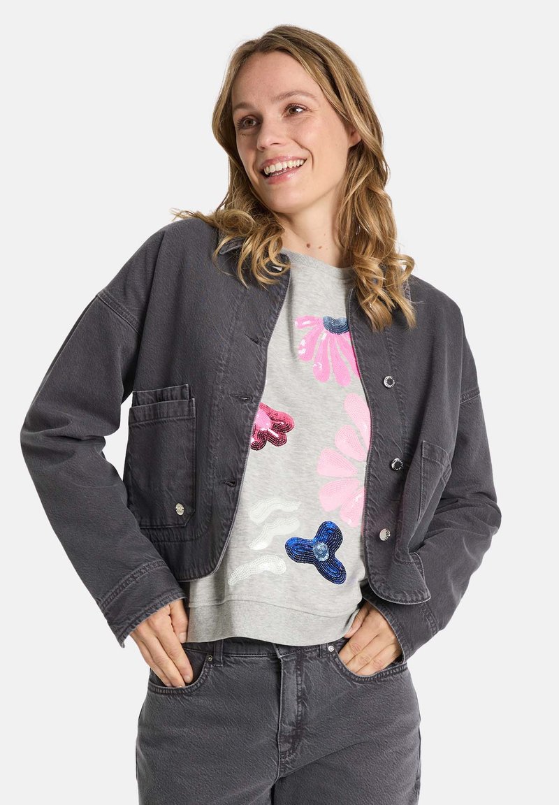 Graue cropped Jacke mit großen Taschen, Knopfverschluss und strukturiertem Stoff, getragen über einem hellgrauen Sweatshirt mit bunten Blumen-Pailletten.