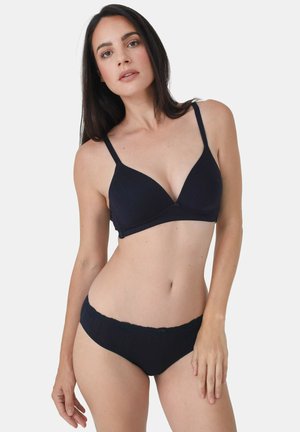 Frau mit langen dunklen Haaren, die einen schwarzen BH mit dünnen Trägern und passende Hipster-Unterwäsche vor einem schlichten hellen Hintergrund trägt.