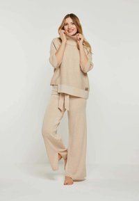 Beige gestrickter Pullover mit hohem Kragen und weiten Ärmeln, kombiniert mit passenden weiten Hosen. Das Material hat ein weiches, strukturiertes Erscheinungsbild.