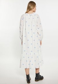 Witte katoenen jurk met blauwe bloemenpatronen, lange mouwen en een gelaagde rok. Beschikt over een geplooide halslijn en een knoopdetail op de rug.