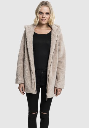 Blonde Frau trägt eine beige flauschige Kapuzenjacke über einem schwarzen Oberteil und zerissene schwarze Hosen, steht vor einem einfarbigen Hintergrund.